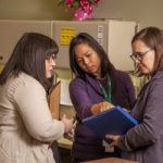 Foto de tres empleadas de cuidados paliativos en un entorno médico, mirando juntos un portapapeles.