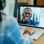 Mujer en un escritorio usando una computadora portátil Surface para hacer una videollamada de Microsoft Teams con un hombre sonriendo y usando un auricular.  El dispositivo de conferencia / reunión de Business Voice está en segundo plano.