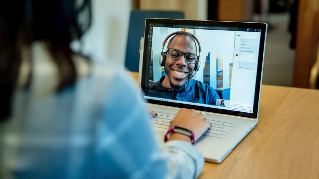 Mujer en un escritorio usando una computadora portátil Surface para hacer una videollamada de Microsoft Teams con un hombre sonriendo y usando un auricular.  El dispositivo de conferencia / reunión de Business Voice está en segundo plano.