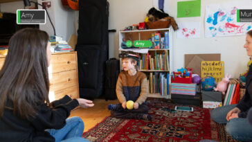 Un niño que usa PeopleLens se sienta en el piso de una sala de juegos con una pelota de tenis ciega en sus manos.  Su atención se dirige hacia una mujer sentada en el suelo frente a él con las manos extendidas.  PeopleLens parece unas pequeñas gafas que se colocan en la frente.  La imagen está marcada con anotaciones visuales para indicar lo que ve PeopleLens y qué sonidos se escuchan.