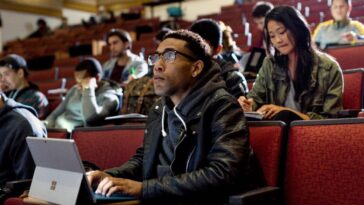 Primer plano de un estudiante universitario masculino sentado en la sala de conferencias de la universidad, mirando hacia adelante mientras toma notas en un Surface Pro.  Otros estudiantes, hombres y mujeres, se sientan detrás de él y toman notas.