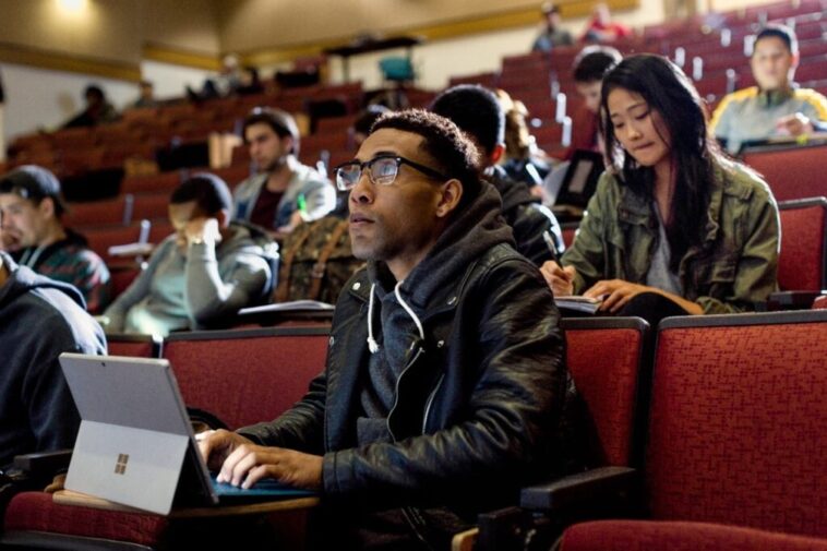 Primer plano de un estudiante universitario masculino sentado en la sala de conferencias de la universidad, mirando hacia adelante mientras toma notas en un Surface Pro.  Otros estudiantes, hombres y mujeres, se sientan detrás de él y toman notas.