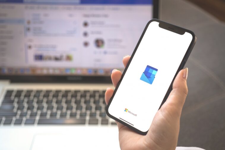 Logotipo de Outlook en la pantalla del teléfono móvil y persona que usa la aplicación Microsoft Outlook en una computadora portátil.