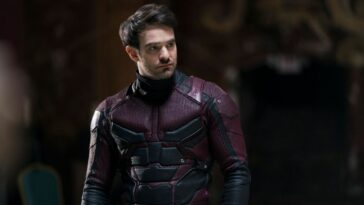 An unmasked Matt Murdock stands in a dark room with his Daredevil suit on in the character