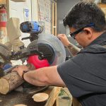 Jimmy, con equipo de protección, está trabajando en un taller de carpintería y cortando un tronco de madera.