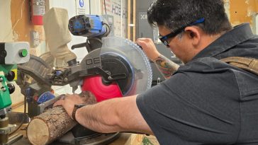 Jimmy, con equipo de protección, está trabajando en un taller de carpintería y cortando un tronco de madera.