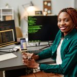 Retrato del desarrollador de TI femenino mirando la cámara y sonreír contra el código de programación en la pantalla de la computadora en el interior de la oficina.