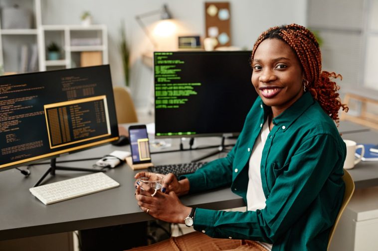 Retrato del desarrollador de TI femenino mirando la cámara y sonreír contra el código de programación en la pantalla de la computadora en el interior de la oficina.
